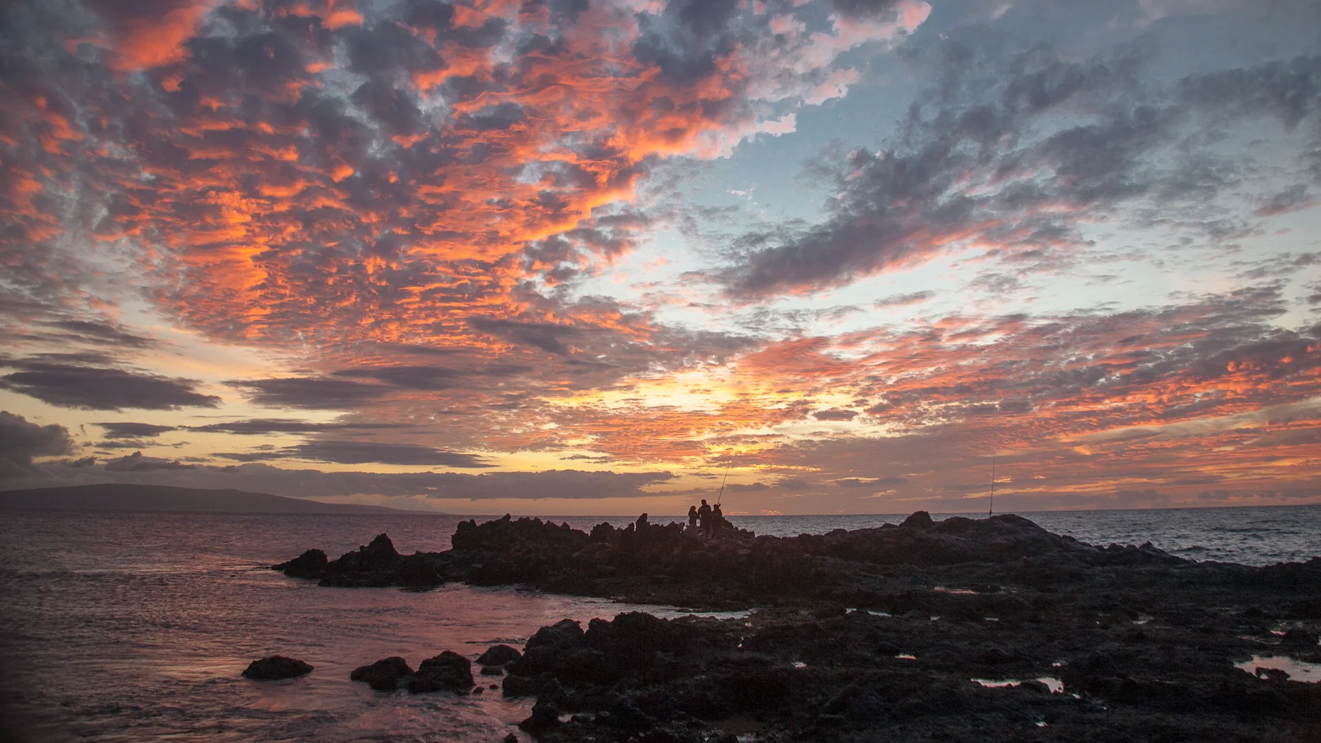 A fiery South Maui sunset over the ocean, silhouetted lava rocks and two anglers at the water's edge near Kihei